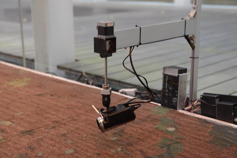Camera attached to a robotic arm. It captures video footage as it slowly pans across the carpet, incorporating the carpet as landscape, just like the ruins.